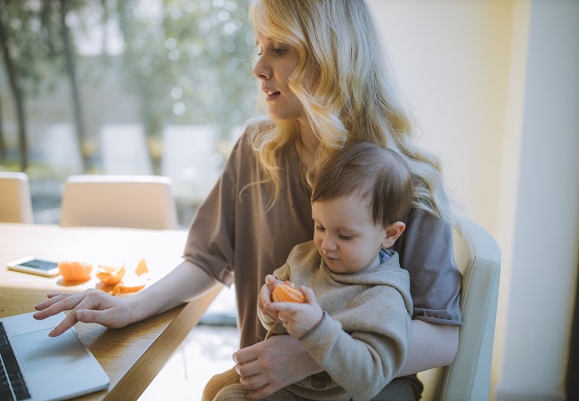 Mama ce lucreaza pe laptop in timp ce isi tine copilul in brate