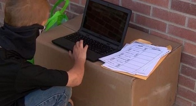Baietel purtand masca de protectie neagra si incercand sa participe la cursuri online cu ajutorul unui laptop sprijinit pe o cutie de carton