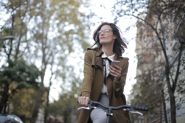 femeie-tânără-cu-ochelari-ce-poarta-o-haina-lunga-maro-si-care-merge-pe-bicicleta-in-timp-ce-intr-o-mână-îsi-tine-telefonul