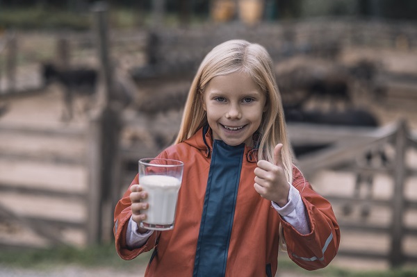 fetita blonda bucuroasa tinand in mana un pahar cu lapte in timp ce in spatele ei se vad animale dintr-o ferma