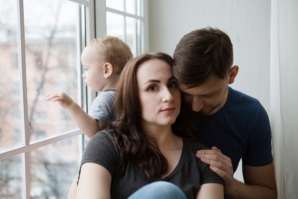mama deprimata incurajata de partenerul ei in timp ce copilul lor se afla in spatele lor si se uita pe fereastra