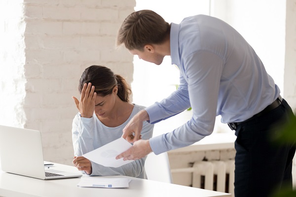 femeie trista la birou in timp ce seful ei nemultumit îi face observatii in legatura cu un document