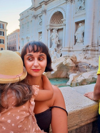 femeie ce isi tine in brate fetita in timp ce se afla cu spatele la una dintre fantanile din Roma