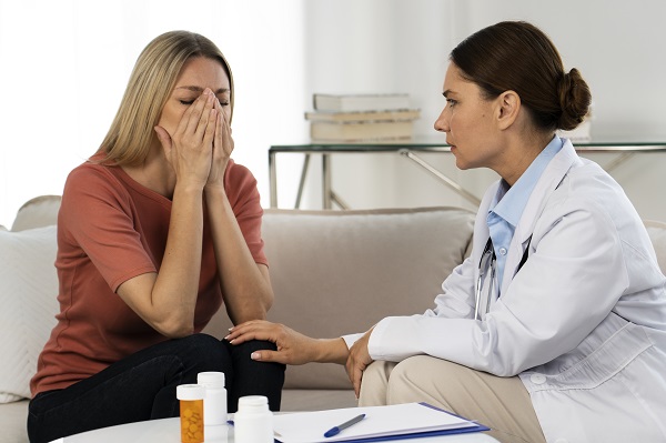 femeie tanara, coplesita, stand pe canapea, in timp ce o doctorita incearca sa o consoleze si sa-i recomande un tratament