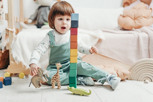 copil in salopeta, stand pe covor si jucandu-se construind un turn din cuburi de lemn colorate