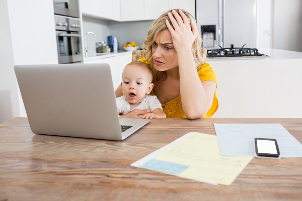 mama stresata, care isi tine bebelusul in brate in timp ce lucreaza pe laptop