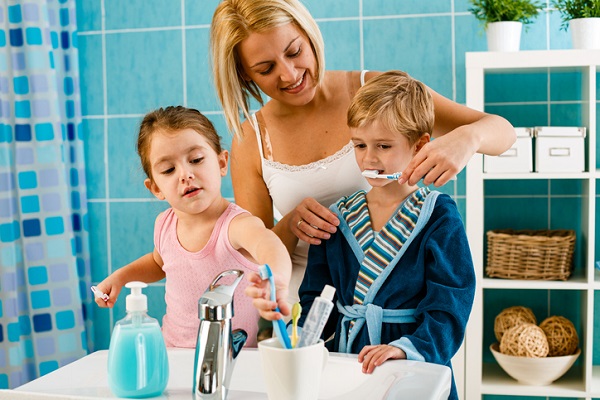 mama care se afla in baie alaturi de copiii ei si isi ajuta baietelul sa se spele pe dinti