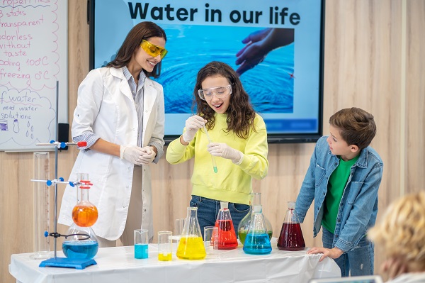 doi elevi care stau alaturi de profesoara entuziasmata, care le ofera explicatii prin intermediul unui experiment la chimie