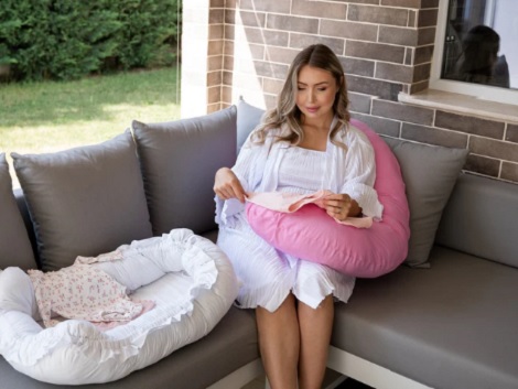 femeie tanara stand pe o canapea pe veranda relaxandu-se pe o perna pentru gravide roz si avand langa ea un cuib pentru bebelusi alb cu volanase