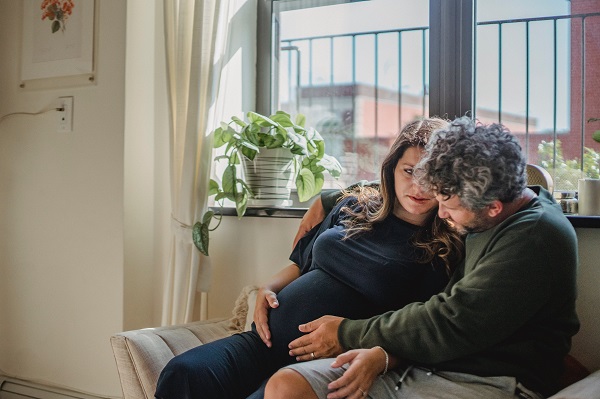 femeie insarcinata stand pe canapea alaturi de partenerul ei care îi atinge afectuos burtica de gravida