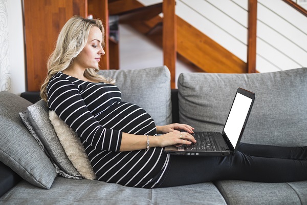 femeie insarcinata care sta intinsa pe canapea si lucreaza pe laptop