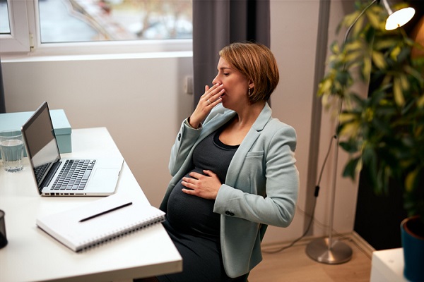 femeie insarcinata obosita, stand la birou in fata laptopului