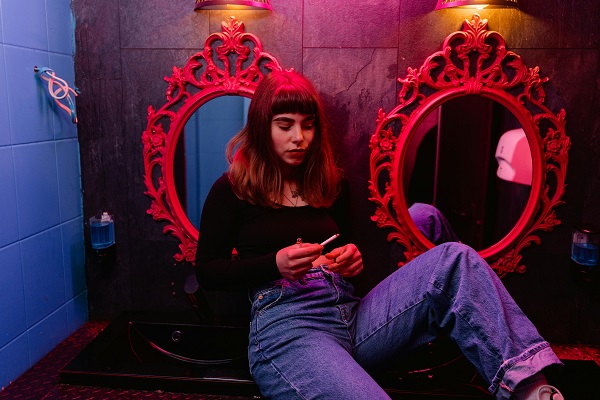 femeie tanara in blugi stand cu spatele la oglinzi rosii in toaleta si uitandu-se la o tigara