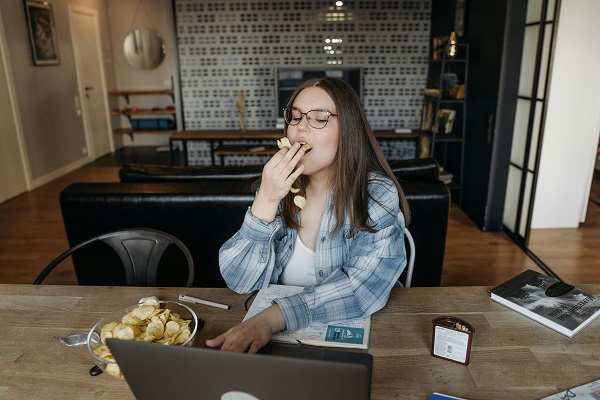 femeie tanara cu ochelari care lucreaza pe laptop si mananca chips-uri
