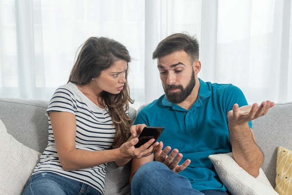 femeie tanara care sta pe canapea langa sotul ei si ii arata ceva pe telefon, in timp ce acesta incearca sa vina cu explicatii