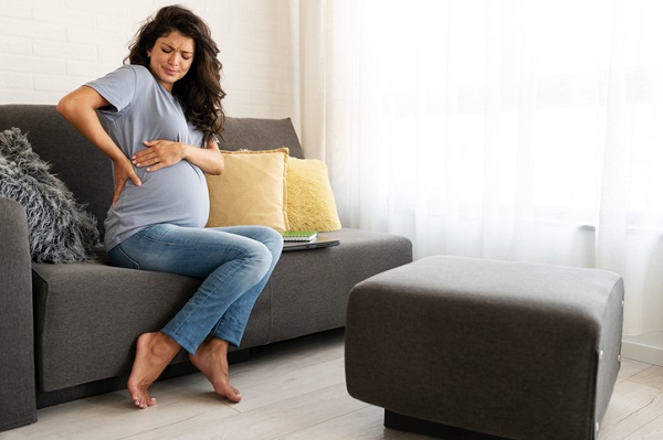 femeie insarcinata stand pe canapea si tinandu-si mainile pe burtica de gravida din pricina disconfortului abdominal