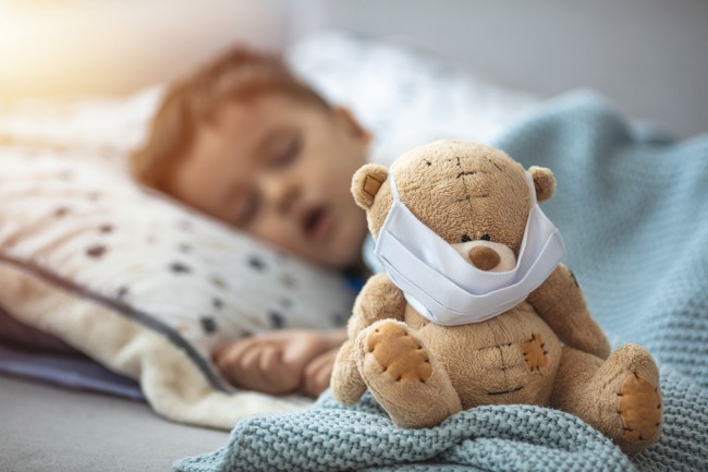 copil care doarme langa un ursulet de plus cu masca de protectie