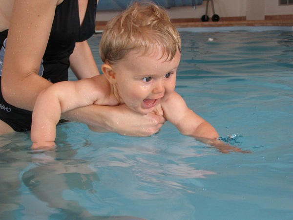 Femeie in costum de baie care a intrat in piscina si isi tine bebelusul care se joaca