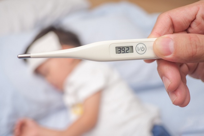 Copil cu febra mare care sta in pat si parinte care ii verifica temperatura