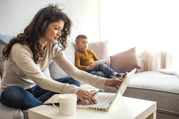 mama-care-lucreaza-la-laptop-in-timp-ce-baietelul-ei-sta-pe-canapea-cu-tableta