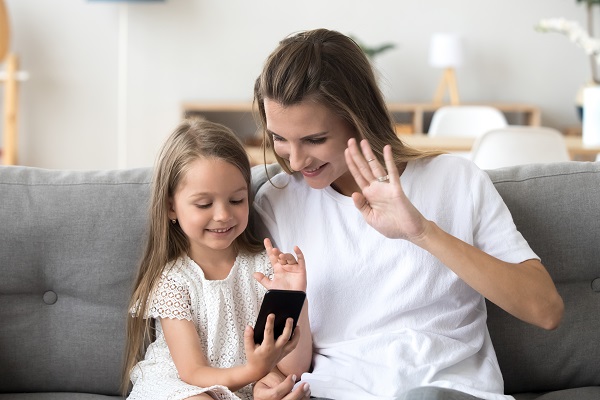 mama-si-fetita-ce-stau-pe-o-canapea-fetita-zambeste-in-timp-ce-utilizeaza-telefonul-iar-mama-ei-saluta-in-timpul-unui-apel-video