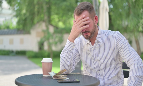 barbat cu barba stand trist cu fata in maini la o masuta in aer liber cu o cafea