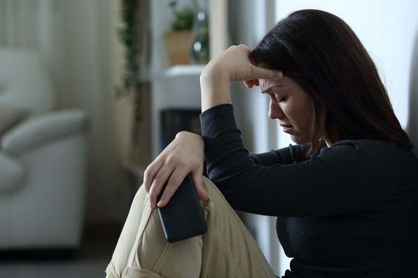 femeie tanara trista rezemandu-se de un perete si tinandu-si telefonul in mana 