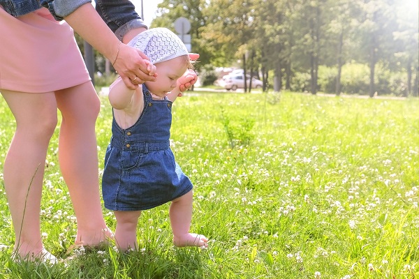 fetita in sarafan de blugi care invata sa mearga pe iarba in timp ce mama ei o tine de manute