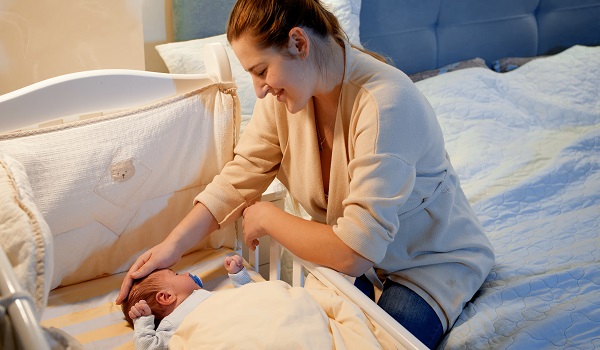 mama zambitoare care sta pe un pat si isi mangaie pe cap bebelusul ce doarme in patutul sau