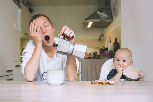 mama epuizata care sta la masa langa bebelusul ei si se străduieste să-si toarne cafea in cana