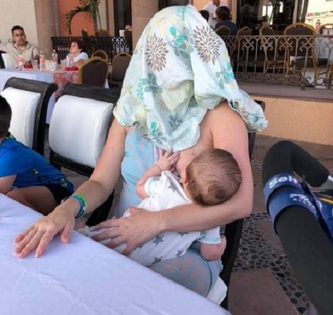 femeie care si-a acoperit capul in timp ce isi alapteaza bebelusul la o masa pe terasa unui restaurant