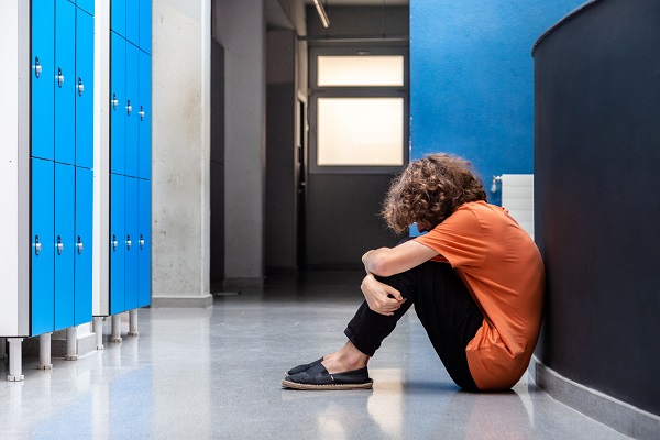 adolescent cu tricou portocaliu si cu parul cret stand ghemuit si deprimat pe holurile scolii
