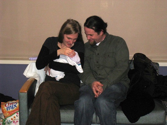 femeie fericita tinand in brate un bebelus, in timp ce alaturi de ea se afla partenerul ei