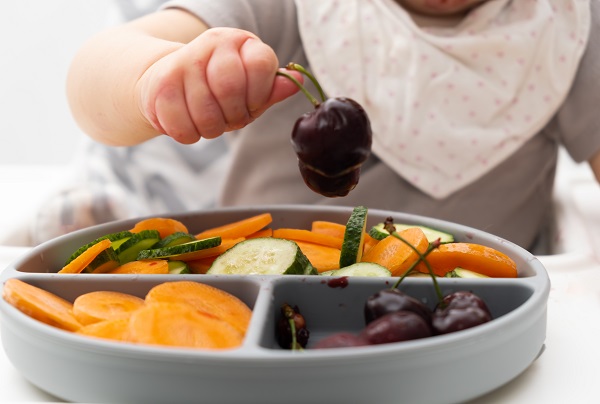 bebelus cu bavetica ce ia in manuta cirese dintr-un vas cu fructe si legume aflat in fata sa