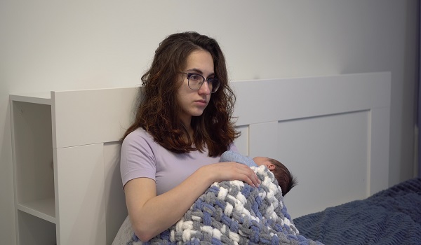 mama tanara cu ochelari stand in pat trista si tinandu-si in brate bebelusul nou-nascut