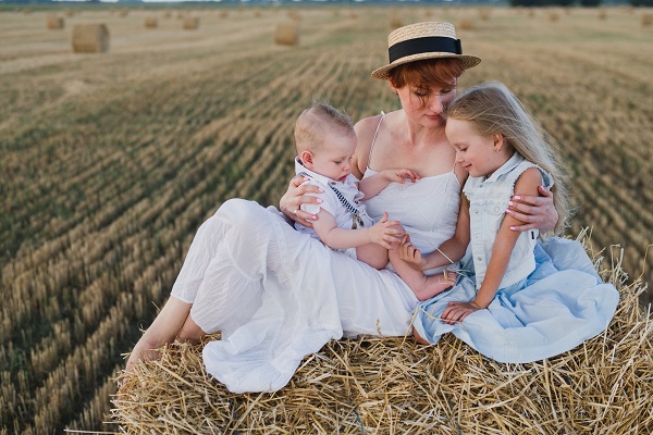 mama imbracata in rochie alba petrecandu-si timpul cu cei doi copii ai ei pe un camp cu baloti de fân