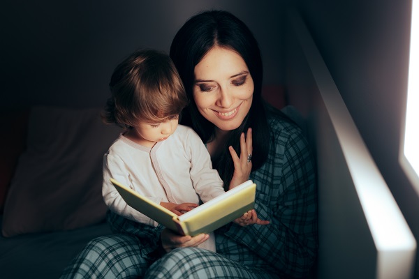 mama vesela in pijama tinandu-si pe genunchi fetita si citindu-i inainte de culcare