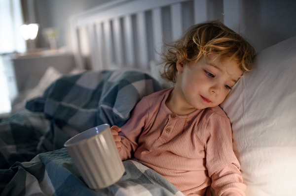 fetita in pijama roz care se trezeste noaptea si sta in pat tinand in mâna o cana de ceai