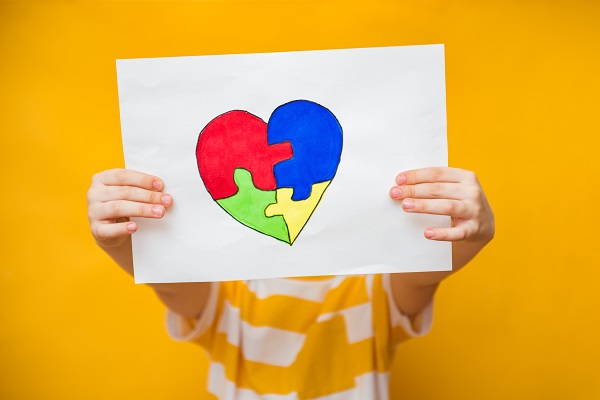 copil cu tricou in dungi albe si galbene tinand in maini o coala de hartie pe care este desenata o inima pentru constientizarea problemelor ce tin de tulburarile de spectru autist
