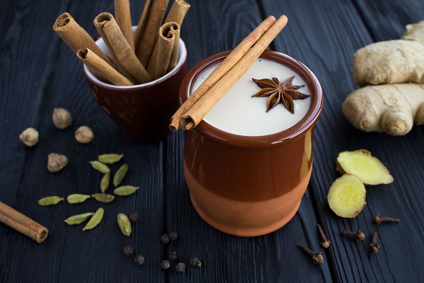 Masala Chai in cana de ceramica, alaturi de care se afla diverse condimente: ghimbir, cardamom, batoane de scortisoara, cuisoare, piper negru