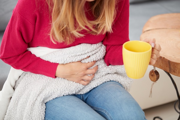 femeie tanara in blugi, fixand cu o mana o patura asezata in zona abdomenului si cu cealalta mana tinand o cana cu ceai