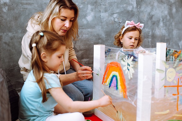 femeie care ajuta doua fetite sa picteze curcubeu in timpul unei activitati artistice