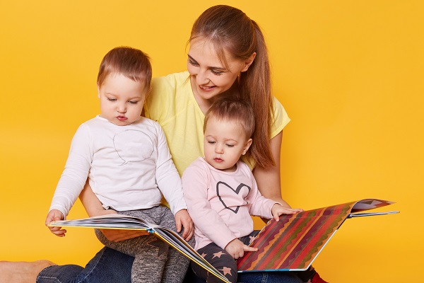 mama fericita care isi tine pe genunchi fetitele gemene si se uita impreuna pe carti