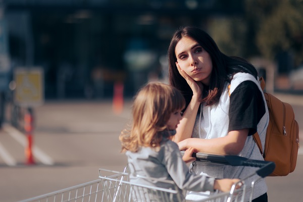 femeie tânara care sta pe ganduri in timp ce impinge cosul de cumparaturi in care se afla o fetita