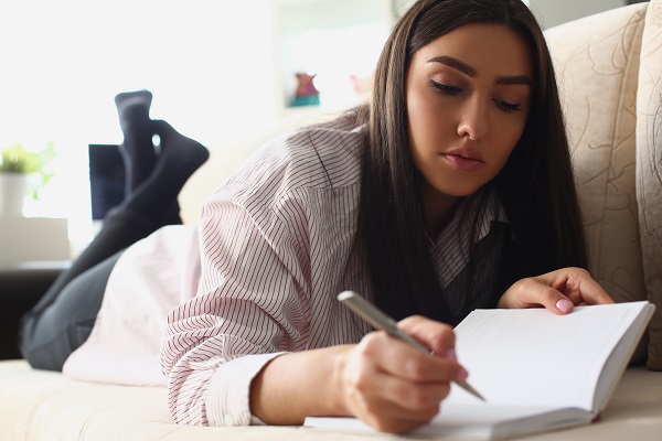 femeie tanara scriind intr-un jurnal in timp ce sta intinsa pe canapea