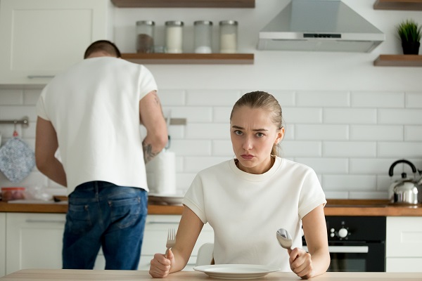 femeie tanara furioasa stand la masa din bucatarie in fata farfuriei goale in timp ce partenerul ei gateste