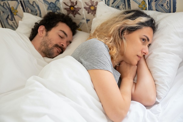 femeie care sta ingandurata in pat in timp ce partenerul ei doarme