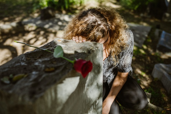 femeie cu parul ondulat stand cu un trandafir rosu si plangand deasupra unui mormant
