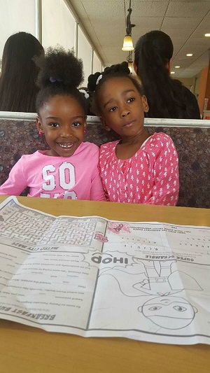 doua fetite de culoare vesele, imbracate in roz, stand la masa la un restaurant