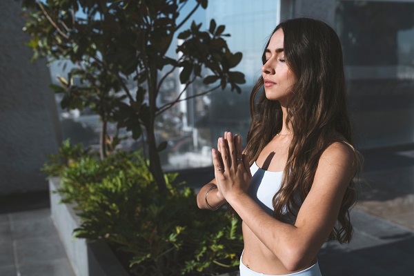 femeie tanara cu parul lung meditand cu ochii inchisi pe terasa unei cladiri